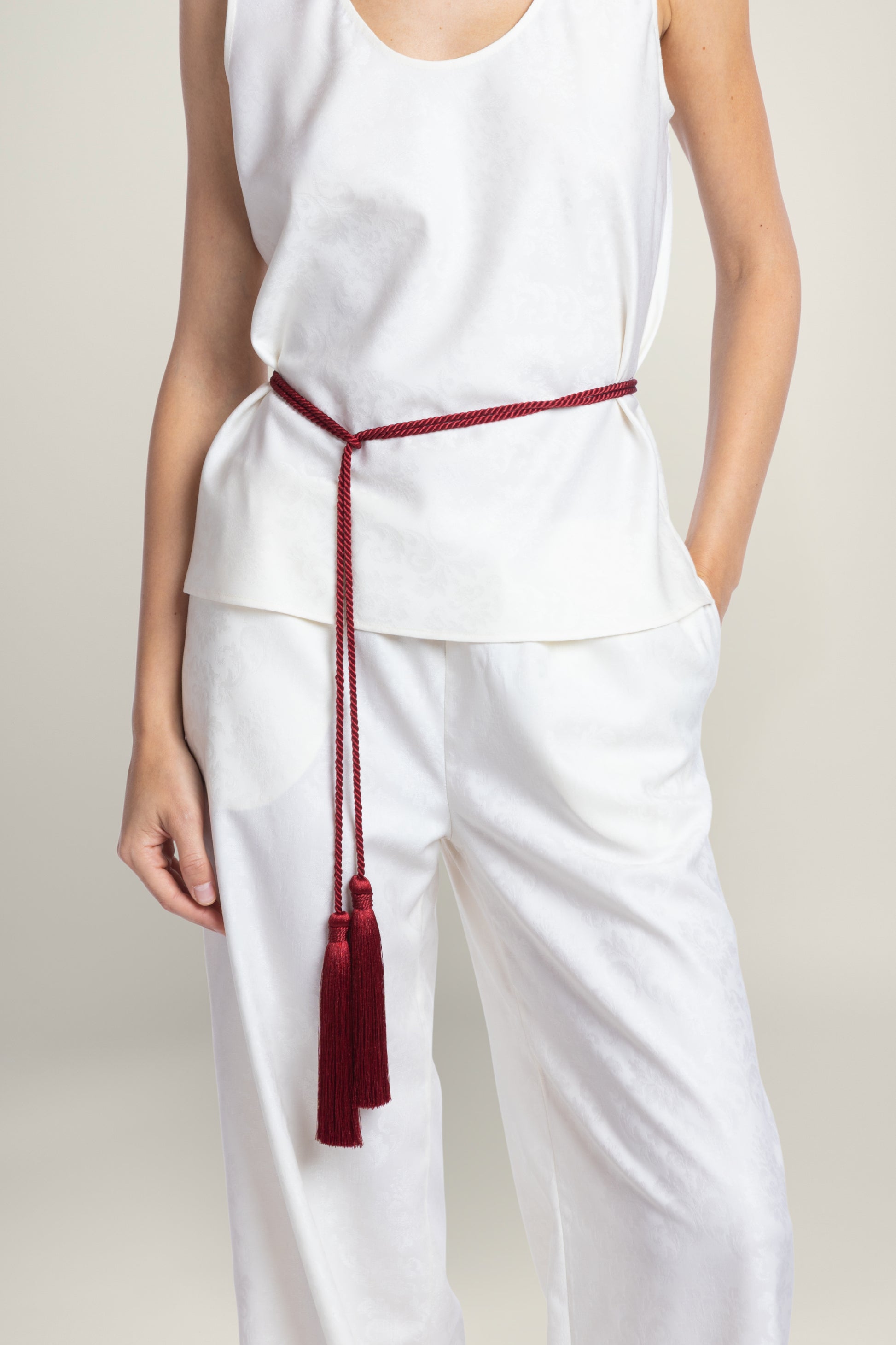 A model wearing a white sleeveless top and matching white wide-leg pants, styled with a dark red cord belt tied at the waist with a tassel.